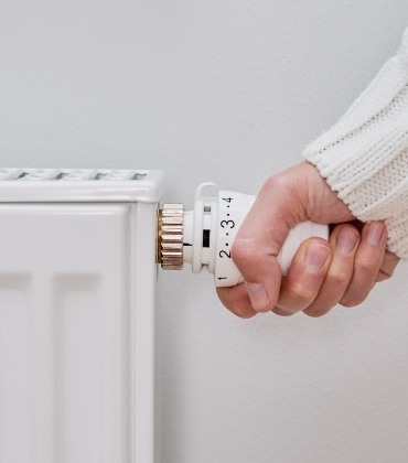 Iemand pakt met 1 hand de thermostaatknop van de radiator vast, om de verwarming hoger of lager te kunnen zetten.