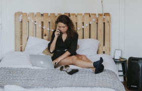 Een vrouw zit met de laptop opengeklapt op het bed, ze heeft een telefoon aan haar oor.