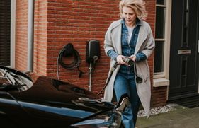 Een vrouw sluit haar elektrische auto aan op de laadpaal, die aan de gevel van het woonhuis hangt.