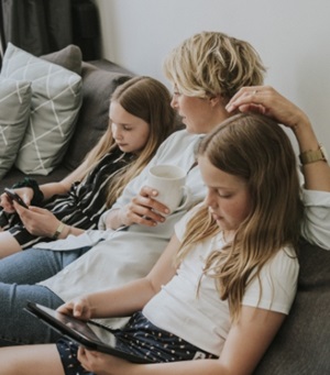 Een vrouw zit op de bank tussen haar beide dochters in. Ze heeft een witte mok vast, en de dochters kijken op hun mobiele telefoons.