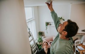 Een man staat op een trapje en hangt een rookmelder op aan het plafond.