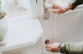 Een persoon houdt met 1 hand de klink van een witte buitendeur vast, en draait met de andere hand de sleutel om in het slot.
