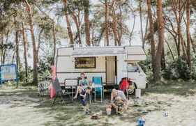 Gezin is aan het kamperen, vrouw zit een tijdschrift te lezen, man en kinderen spelen voor de camper