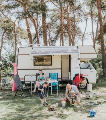 Gezin is aan het kamperen, vrouw zit een tijdschrift te lezen, man en kinderen spelen voor de camper