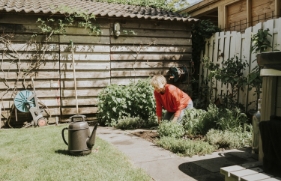 Een vrouw met blond haar werkt in de tuin. Ze zit op haar knieën en harkt de tuingrond los. Naast haar staan diverse soorten tuingereedschap.