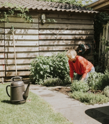 Een vrouw met blond haar werkt in de tuin. Ze zit op haar knieën en harkt de tuingrond los. Naast haar staan diverse soorten tuingereedschap.