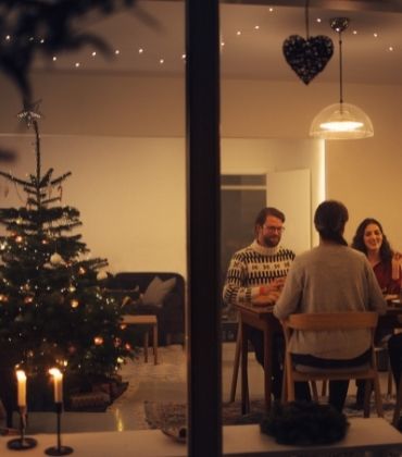 4 mensen zitten binnenshuis aan een keukentafel, naast een rijkversierde kerstboom.