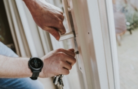 De handen van een persoon; de linkerhand pakt een deurklink vast, de rechterhand steekt een sleutel in het slot daar direct onder.