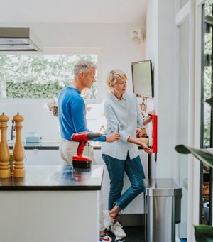 Een man en een vrouw bevestigen samen een blusdeken aan de muur in de keuken.