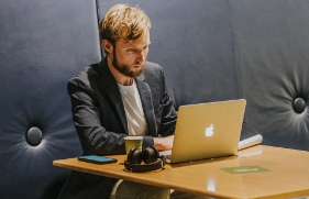 Een man werkt op zijn laptop, die op een tafeltje voor hem staat. Naast de laptop ligt ook zijn smartphone op het tafelblad.