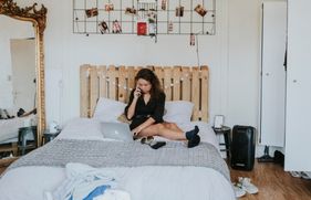 Jonge vrouw zit op het bed in haar studentenkamer en belt terwijl ze op de laptop bezig is.