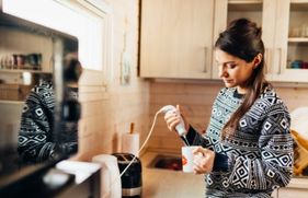Vrouw in keuken met staafmixer. Lees hoe je de kans op kortsluiting en elektriciteitsschade in huis verkleint.